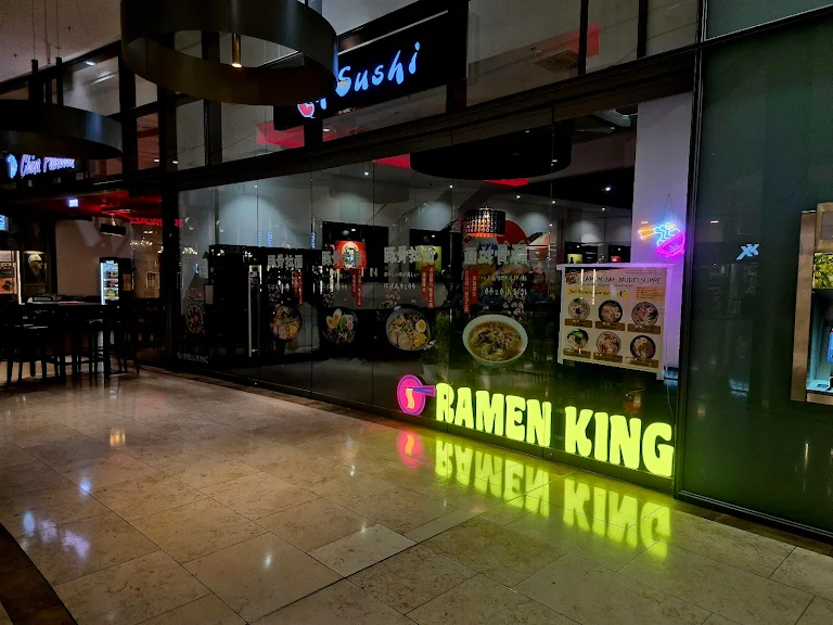 Außenansicht eines Restaurants mit dem Namen "Ramen King". Die Glasfront zeigt einen beleuchteten gelben Schriftzug. Daneben hängt ein Poster mit Bildern von Speisen. Im Inneren sind Tische und Stühle zu erkennen, und über der Fassade leuchtet ein weiteres Geschäftsschild mit dem Wort "Sushi". Die Umgebung wirkt modern und einladend.