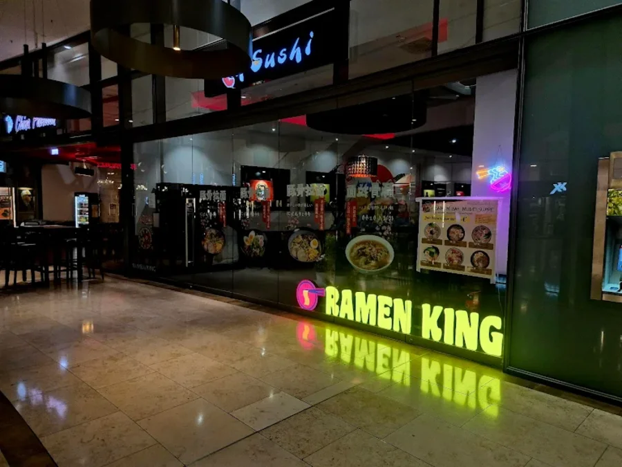 Außenansicht eines Restaurants mit dem Namen "Ramen King". Die Glasfront zeigt einen beleuchteten gelben Schriftzug. Daneben hängt ein Poster mit Bildern von Speisen. Im Inneren sind Tische und Stühle zu erkennen, und über der Fassade leuchtet ein weiteres Geschäftsschild mit dem Wort "Sushi". Die Umgebung wirkt modern und einladend.
