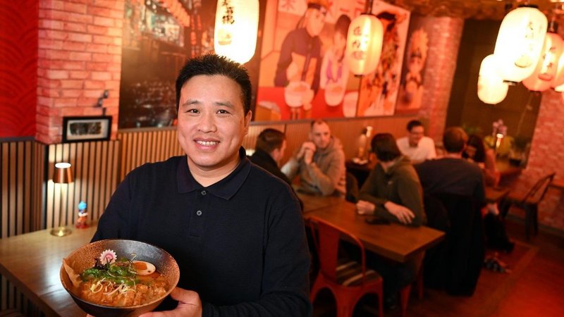 Ein Mann steht lächelnd in einem gemütlich beleuchteten asiatischen Restaurant und hält eine große Schüssel Ramen in den Händen. Im Hintergrund sitzen mehrere Gäste an Holztischen, die Wände sind mit Bildern und Laternen im asiatischen Stil dekoriert.